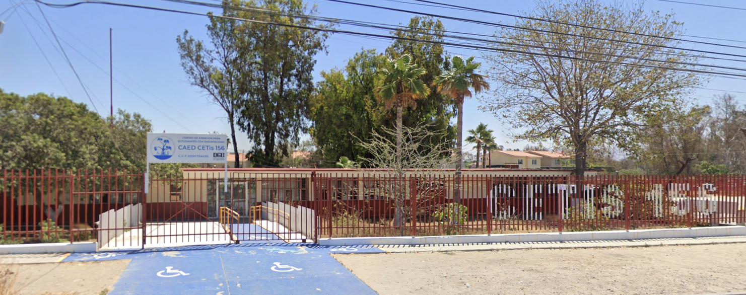 En la fotografía se muestra una reja roja que rodea el plantel, encima de ella se encuentra un cartel donde dice CAED CETIS 156, del lado izquierdo se encuentran las rampas de acceso.